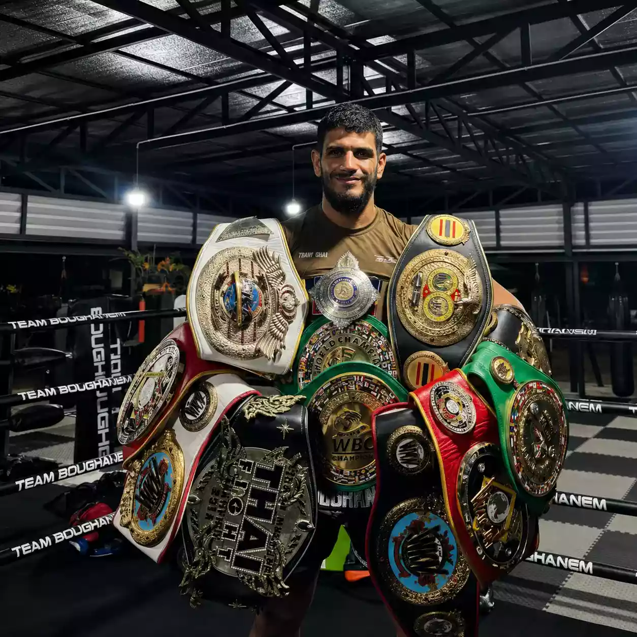 Youssef Boughanem, légende du Muay Thaï, posant fièrement avec ses nombreuses ceintures de champion du monde dans sa salle de boxe
